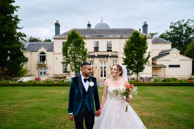 A bride and groom walking hand-in-hand towards the camera with a large, cream house behind them